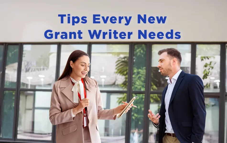 Two business professionals stand outside a modern office building having a discussion. The woman holds a clipboard and smiles while speaking to the man, who listens attentively. Text above them reads, “Tips Every New Grant Writer Needs.”
