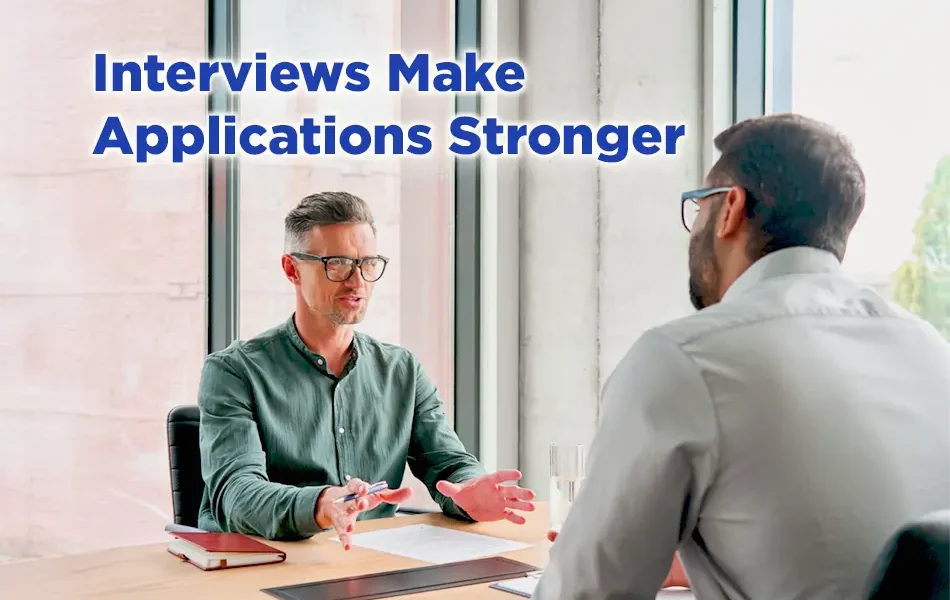 A job interview scene with two men seated across a desk in a bright office. One man gestures while speaking, and the text “Interviews Make Applications Stronger” appears above them.