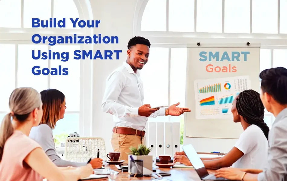 A diverse team sits around a conference table while a presenter gestures toward a flip chart labeled ‘SMART Goals’ with charts and graphs. Text on the image reads ‘Build Your Organization Using SMART Goals’ in a bright, modern meeting space.