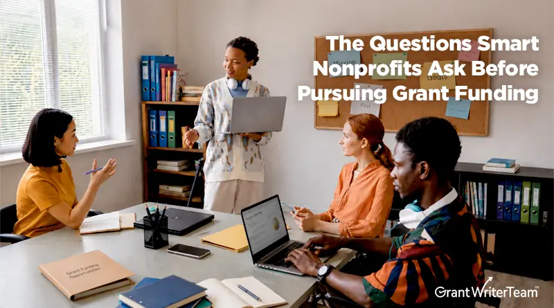 A diverse group of nonprofit professionals meets in an office conference room while discussing strategy and funding goals. One team member stands and presents using a laptop while others listen and take notes around a table with computers, folders, and documents. Text overlay reads, "The Questions Smart Nonprofits Ask Before Pursuing Grant Funding," with the GrantWriterTeam logo in the corner.
